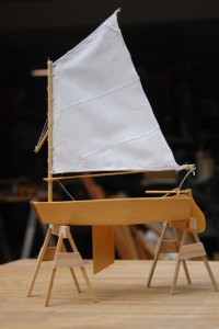 Optimist Pram model on model sawhorses.  Centerboard, rudder, and sail are in profile in this side view.